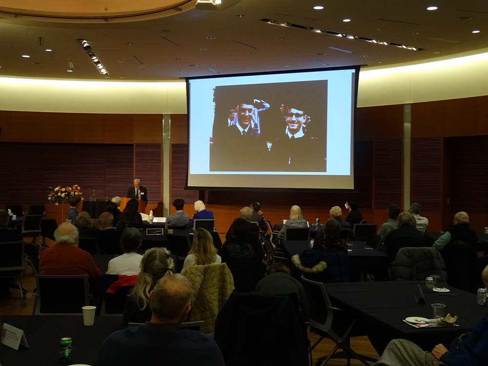 A video screen shows an image of John Kutzbach wearing a cap and gown at a graduation ceremony