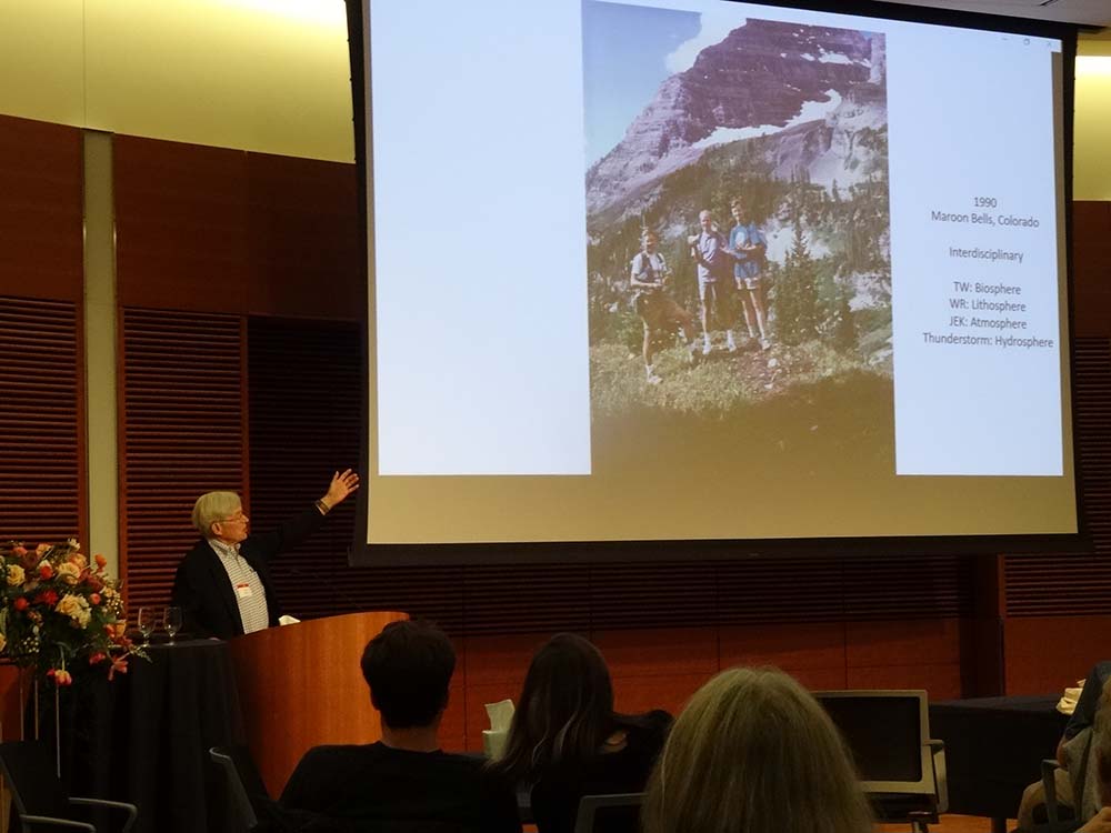A video screen shows an image from 1990 of John Kutzbach standing on a mountain in Maroon Bells, Colorado