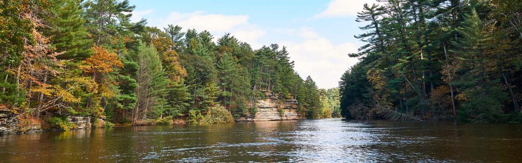 A river through Upham Woods with shores lined with pine trees and rock formations